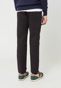 Persoon in zwarte broek, groene en beige sneakers, en een marineblauwe trui, staande tegen een effen lichte achtergrond met de rug naar ons gekeerd.