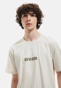 Camiseta de algodón crema con mangas cortas, cuello redondo y diseño impreso en marrón con la palabra "sueño" en la parte frontal. Textura suave y ligera.