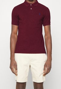 Man i maroon kortärmad polo-tröja och beigefärgade knälånga shorts, stående mot en enkel ljus bakgrund.