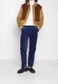 Hnědá manšestrová bunda na zapínání na knoflíky, bílá texturovaná košile, modré manšestrové kalhoty a černé boty s tlustou podrážkou.