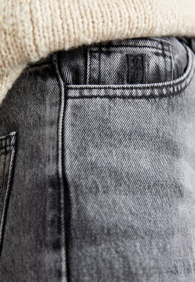 Black denim jeans featuring a textured finish, visible stitching, and a small brand label on the back pocket, partially covered by a light sweater.