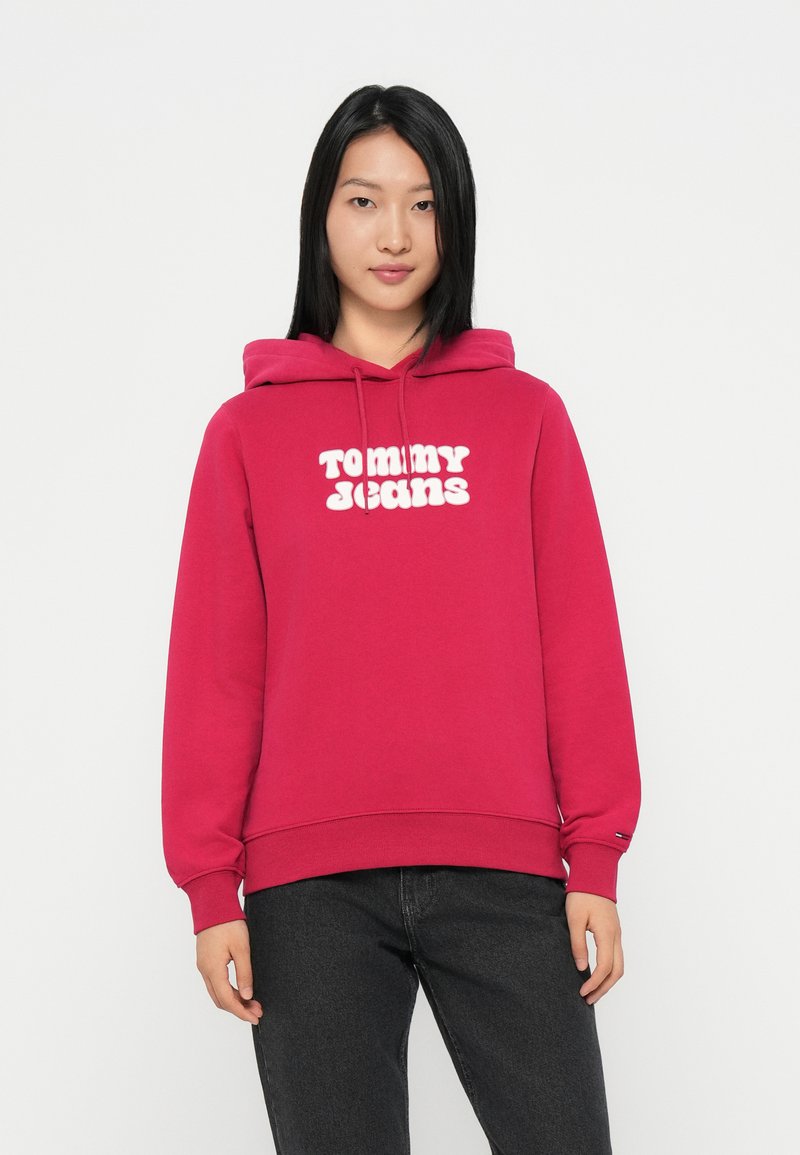 Sweatshirt à capuche fuchsia en mélange de coton. Présente le logo "TOMMY JEANS" en blanc. Manches et ourlet côtelés. Associé à un jean foncé.