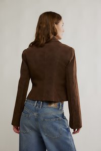 Braune Wildlederjacke in Kurzform mit langen Ärmeln, spitzem Kragen und figurbetonter Silhouette, kombiniert mit hoch taillierten Jeans.