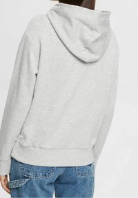 Felpa grigia con cappuccio, caratterizzata da una texture morbida, polsini e vita a costine, e una vestibilità rilassata. Indossata con jeans di denim blu.
