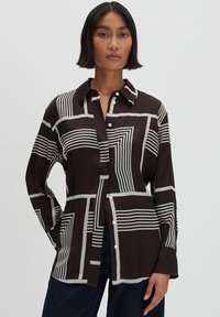 Camicia a maniche lunghe con bottoni di colore marrone scuro, decorata con motivi geometrici bianchi. Presenta un colletto e una vestibilità rilassata con spacco laterale.