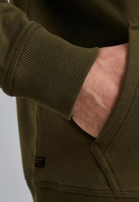 Olijfgroene sweatshirtstof heeft ribgebreide manchetten en een zijzak, en toont textuur en stikseldetails.