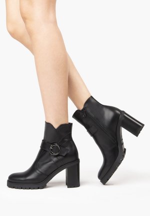 Botas de tobillo de cuero negro con tacón grueso, correa decorativa y acento de anillo, borde superior con volantes y detalle de suela texturizada.