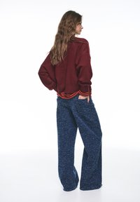 Tummanpunainen yli-koon neulepaita, jossa on teksturoitu pinta ja raidallinen kaulus, yhdistettynä löysiin, sinisiin pilkullisiin housuihin. Molemmat luovat rentoutuneen siluetin.