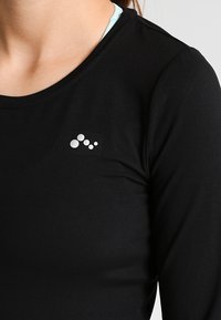Schwarzes Langarmshirt aus glattem Stoff, mit Rundhalsausschnitt und einem kleinen silbernen Logo oben links.