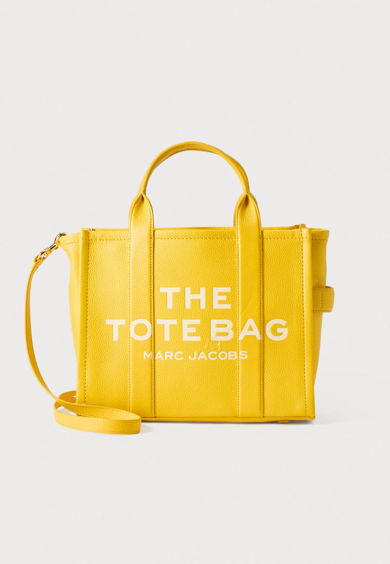 Svetlo rumena usnjena torba s kratkimi ročaji, odstranljivim ramenskim trakom in natisnjeno oznako "THE TOTE BAG MARC JACOBS" spredaj.