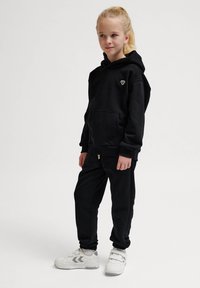 Schwarzer Hoodie mit Fronttasche und Logo, kombiniert mit passenden schwarzen Jogginghosen. Das Modell trägt weiße Sneakers mit grauen Akzenten und steht selbstbewusst.