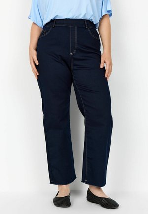 Person trägt dunkelblaue Jeans mit geradem Bein und rohen Säumen, ein hellblaues, lockeres Hemd und schwarze gewebte Ballerinas, steht an einer weißen Wand.