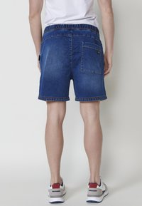 Tumši zili džinsu šorti ar elastīgu jostasvietu, aizmugures kabatu un šūtām apmalēm, apvienoti ar gaišas krāsas sporta apaviem.