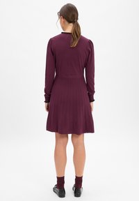 Sukienka dzianinowa w głębokim burgundzie, z długimi rękawami, dopasowanym górnym partem i plisowanym dołem. Czarna ozdoba przy dekolcie i pasujące skarpety.