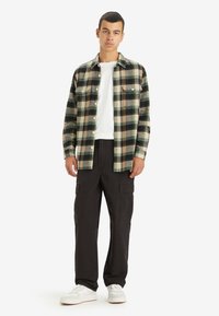 Grøn, sort og beige ternet flannel skjorte med knaplukning; båret over en hvid t-shirt, parret med sorte cargo-bukser og hvide sneakers.
