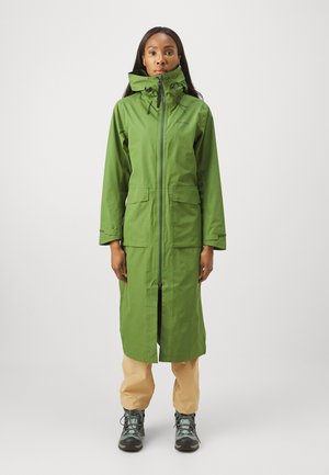 Abrigo largo impermeable verde con cuello alto, cierre de cremallera, dos bolsillos frontales y cordones ajustables; usado sobre pantalones beige y botas.