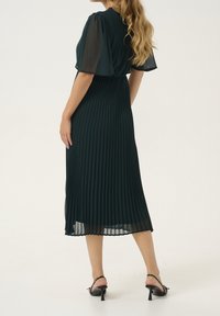 Tmavě zelené plisované midi šaty s průhlednými rukávy, zúženým pasem a volným střihem. Kombinováno s černými sandály na podpatku.