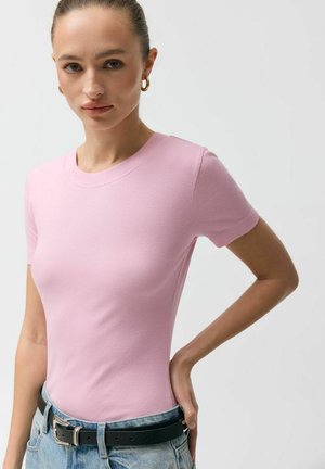 Młoda kobieta związała włosy, nosi złote kolczyki koła, dopasowany różowy top z krótkim rękawem, jasnoniebieskie dżinsy i czarny pasek, stoi z ręką na biodrze.