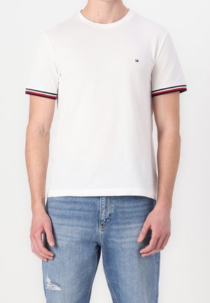 Hombre con camiseta blanca de manga corta con puños a rayas en rojo, blanco y negro y vaqueros azules rotos, de pie frente a fondo blanco.