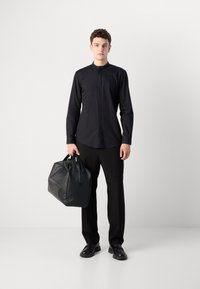 Schwarzes Langarmshirt, elegantes Design mit hohem Kragen, getragen mit schwarzen Hosen und polierten Schuhen, hält eine strukturierte schwarze Tasche.