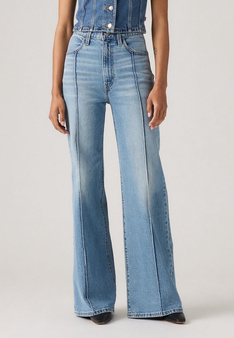 Femme portant un jean taille haute bleu clair à jambes larges avec des coutures verticales à l'avant, associé à un top sans manches en denim et des chaussures noires.