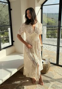 Femme en robe en dentelle crème, pieds nus, debout près d'une fenêtre avec un sol en pierre et un panier, regardant dehors un paysage vallonné et vert.