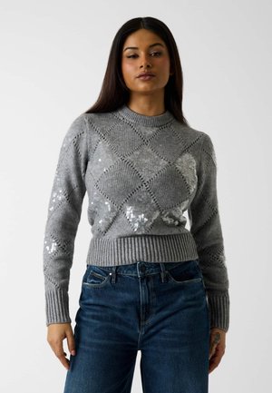 Kobieta w szarym dzianinowym swetrze z diamentowym wzorem i cekinowymi detalami, w połączeniu z wysokim stanem niebieskich dżinsów, stojąca na tle gładkim.