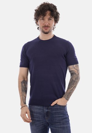 Uomo con capelli ricci che indossa una camicia blu navy a maniche corte e jeans, mostrando tatuaggi dettagliati su entrambi gli avambracci, mano in tasca.