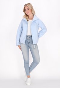 Donna bionda posa con sicurezza indossando una giacca azzurra chiara e una maglietta bianca, abbinate a jeans chiari e scarpe da ginnastica bianche su uno sfondo neutro.