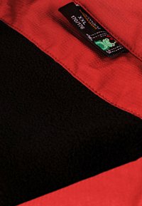 Rote Stoffauskleidung mit schwarzem Fleece-Innenfutter. Verfügt über ein Etikett mit Text und einer kleinen grünen Grafik. Sichtbare Naht entlang der Kante.