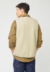 Gilet in pile beige con colletto alto, caratterizzato da una superficie testurizzata, rifiniture verdi a contrasto e indossato sopra una maglietta a maniche lunghe verde oliva.