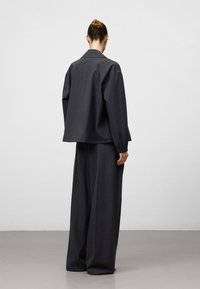 Donkergrijze, oversized jas met een kraag, gecombineerd met losvallende broek. Beide hebben een gestructureerde stof en elastische manchetten.