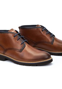 Botas de cuero marrón hasta el tobillo con cordones azules, textura suave, costuras sutiles y suela blanca en contraste. Diseño de punta redondeada.