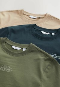 Trois t-shirts en coton empilés. Les couleurs sont beige, vert foncé et marine. Ils ont des cols ronds et des textes imprimés sur les t-shirts vert et marine.