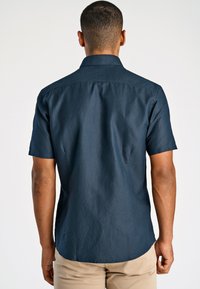 Man die met zijn rug naar de kijker staat en een donkerblauw shirt met korte mouwen en lichtbeige broek draagt, tegen een effen witte achtergrond.
