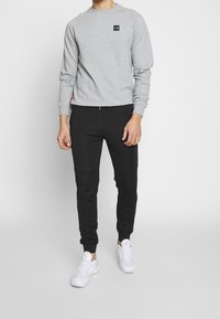 Felpa grigia con un patch logo nero, abbinata a pantaloni neri affusolati con un design testurizzato e sneakers bianche.