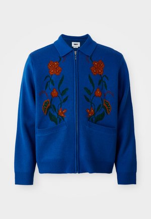 Blauwe gebreide cardigan met een rits, voorzien van bloemenborduurwerk in oranje en groen, en twee voorzakken met ribbels aan de mouwen.