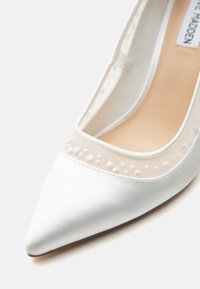 Λευκή γόβα με μυτερή μύτη και διαφανές δίχτυνο πάνελ διακοσμημένο με μικρές λευκές χάντρες κατά μήκος της άκρης, με ορατή μπεζ εσωτερική επένδυση.