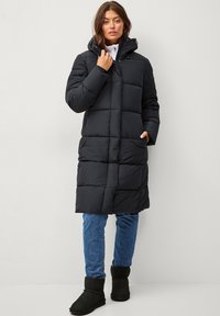 Zwarte gewatteerde lange jas met een capuchon, voorzien van een quiltontwerp en voorzakken, gecombineerd met blauwe jeans en zwarte enkellaarsjes.