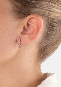 Różowe złote kolczyki w kształcie kółek z delikatnym układem małych, błyszczących kamieni wzdłuż przedniej krawędzi, prezentujące teksturowany wzór.