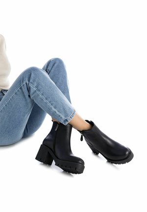 Botas de cuero negras hasta el tobillo con un tacón grueso y suela resistente, con cremallera lateral y textura suave. Combinadas con jeans de un azul claro.