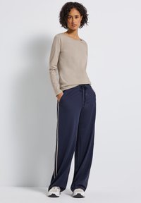 Donna con capelli ricci, che indossa una camicia beige a maniche lunghe, pantaloni larghi blu con strisce laterali e sneakers bianche, in piedi con le mani in tasca.