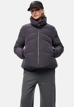 Giovane che indossa una giacca imbottita nera, un cappellino nero con logo e pantaloni grigi strutturati, in piedi con le mani nelle tasche della giacca.
