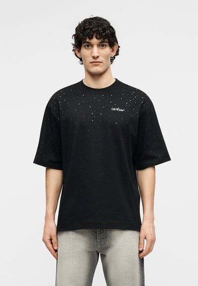 Νέος άνδρας με σκούρα, σγουρά μαλλιά, φοράει μαύρο κοντομάνικο μπλουζάκι με λευκά στίγματα και λογότυπο "Off-White", σε συνδυασμό με ανοιχτό γκρι τζιν.