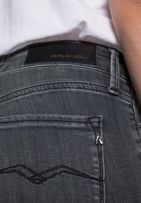 Close-up of dark gray Replay jeans back pocket and waistband worn with white shirt.
