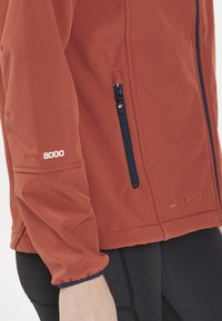 Röd softshell-jacka med marinblå detaljer, ficka med dragkedja och broderad "WPRO 8000" på ärmen. Lättviktig, figursydd design.