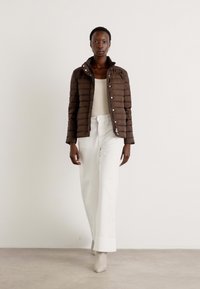 Doudoune marron avec un design matelassé horizontal, haut blanc à manches longues et pantalons blancs évasés. Le modèle porte des bottines beige à la cheville.