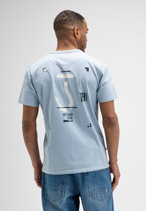Man draagt lichtblauw shirt met korte mouwen met abstracte zwart-witte prints en blauwe spijkerbroek, met de rug naar de camera tegen een effen achtergrond.