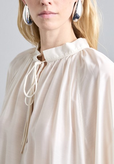 Blusa beige chiaro con colletto alto, dettagli con cordino e tessuto arricciato. Orecchini ovali argento completano l'outfit.