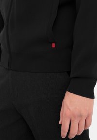 Felpa nera con zip realizzata in un tessuto morbido, con polsini a coste, una tasca e un piccolo logo rosso vicino all'orlo. Pantaloni neri texturizzati.